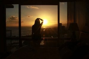 A woman in shadows stands before her floor-to-ceiling bedroom window. Through the window, she watches the golden sun setting.