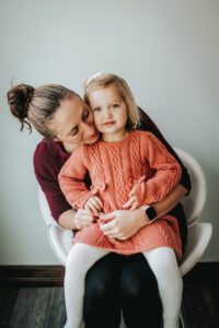 Aubrey, who has SLC6A1, and her mother Erika sit on a chair together. Aubrey has blonde hair, an orange sweater dress, and white leggings.