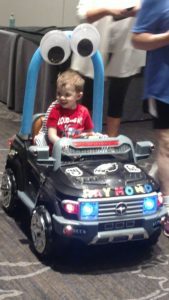 Raymond, who has VAMP2, sits in a toy car. The car is black with red and blue lights. Raymond is wearing a red shirt with white writing.