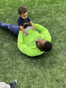 Seattle Seahawks DE Shelby Harris with his son, who has FPIES. Shelby is laying on the ground with his son on his stomach. Shelby is wearing a bright green shirt and jeans. His son is in a black shirt and tan pants.