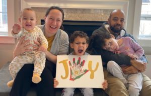 Whitney, Havilah (who has CDKL5), and the rest of the Mitchell family sit around the fireplace. The middle son is holding a sign that says JOY