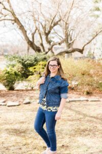 Emily has Stickler syndrome. She wears jeans and a jean jacket and smiles at the camera. She is in front of a tree with no leaves and green bushes.