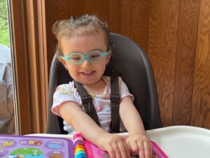 Emma, who has Galloway-Mowat syndrome, sits in her chair. She is wearing green glasses and has her hands stretched out over a colorful toy.