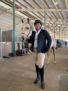Sarah, who has SPS, stands in the stables with one of her horses. She is wearing full riding gear.