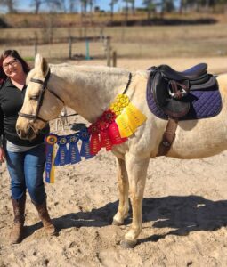 Sarah has SPS and Addison's disease. She stands with her horse who is wearing multiple ribbons and medals.