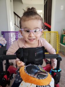 Emma, who has Galloway-Mowat syndrome, sitting in her chair. She is wearing pink glasses and a pink shirt.