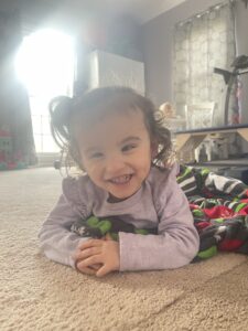 Emma has Galloway-Mowat syndrome. She is laying on the floor in a purple shirt and smiling at the camera.
