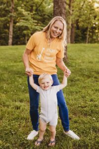Kristin and her son Kayden, who has Chiari malformation. Kristin has long blonde hair and is wearing an orange shirt and genes. She holds Kayden's hands; he is in an all-white outfit. 