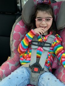 Savannah, who has Ogden syndrome, smiling at the camera. She is buckled into her carseat and is wearing blue pants and a rainbow shirt.