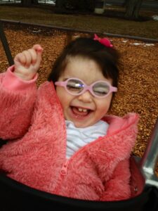 Savannah, who has Ogden syndrome, smiling and waving her hand in the air. She is wearing pink glasses and a pink fuzzy coat over a white shirt.