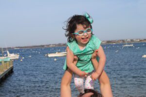 Savannah, who has Ogden syndrome, being lifted into the air on vacation. She has curly brown hair and is wearing a turquoise dress.