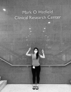 Tracy, who has mastocytosis, standing in front of a wall. She is pointing up towards the words Mark O. Hatching Clinical Research Center.