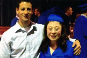 Laura, who has Carpenter syndrome, stands next to a man with a white striped button down shirt. She has just graduated and is wearing bright blue graduation cap and gown.