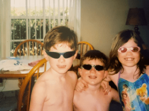 Nicole, who has ALPS, and her brothers. All three children are wearing sunglasses and staring at the camera. Nicole is wearing a blue floral bathing suit.
