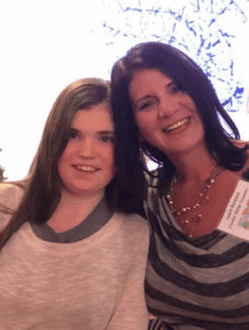 Laura and her daughter Nicole, who has ALPS. Both are smiling at the camera. Nicole is wearing a pink shirt and has long brown hair. Laura has long brown hair, a necklace, and a gray and white striped shirt. 