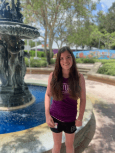 Nicole has ALPS. She stands in front of a fountain with bright blue water. She is wearing a purple shirt and black shorts and is smiling at the camera. Her brown hair falls below her shoulders.