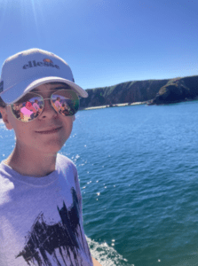 Lachlan, who has spontaneous periodic hypothermia, by the water. He takes a selfie in front of some glistening blue water. He is wearing a white hat, a white shirt with the Batman symbol, and shining sunglasses.