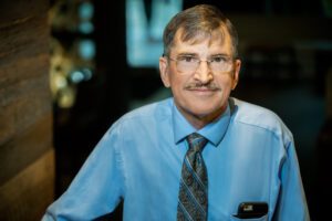Jack, the executive director of FSIG, has dealt with Fabry disease symptoms for his entire life. In this photo, Jack has brown hair, glasses, a mustache, and is wearing a blueshirt with a dark blue striped tie. 