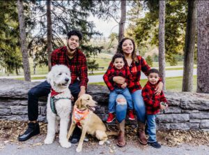 The Cabrera family sits in matching red and black flannel shirts and jeans. They are in front of trees with their two dogs.