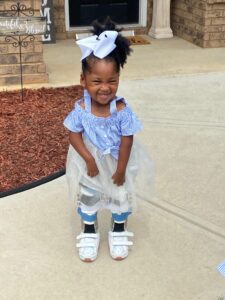 MiKenli, who has AMC, stands on a sidewalk. She has two casts on both legs. She wears a blue shirt, a blue tutu, and a blue bow in her hair.