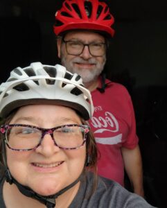 Pam, who has systemic mastocytosis, and her husband. She smiles at the camera. Pam is wearing glasses and a white and black striped biking helmet. Her husband stands behind her. He has silver hair, a red Coca-Cola shirt, and a red biking helmet.