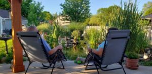 Pam, who has systemic mastocytosis, sits in the backyard with her husband. They are sitting in black chairs and facing a bright green garden.