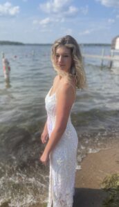 Justice stands in what looks like a white sparkly prom dress. Her blonde hair falls over her shoulder. She is on the beach