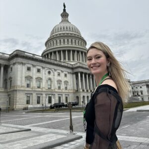Justice, who has PWS, stands in front of the US Capitol Building. She has long blonde hair and a black shirt with semi-sheer sleeves.