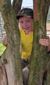 Ryan, who has NPC, sits in a tree smiling at the camera. He's wearing a hat and a yellow shirt