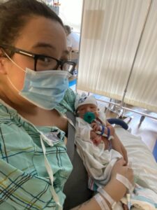 Angela is in the hospital after giving birth to Yiannis, who has IRF2BPL. She is wearing a green gown and a blue mask with black glasses. Yiannis is in her arms with various tubes.