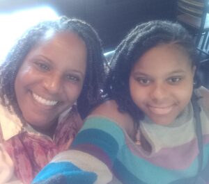 Ronda and her daughter Raeah, who has MCTD, smile at the camera. Both have dark brown hair and pretty smiles. Raeah is wearing a multi-colored sweater.