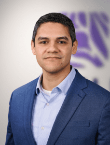 Dan Gallo of Zevra Therapeutics, which is developing a treatment for Niemann-Pick disease type C (NPC), stands in front of the camera. He is smiling at the camera in front of a purple background. Dan wears a light blue buttoned up shirt as well as a dark blue coat jacket.