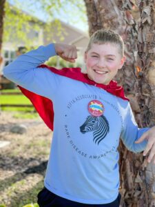 Liam, who has KAND, stands in front of a tree. He is wearing a blue shirt, blue pants, and a red cape.